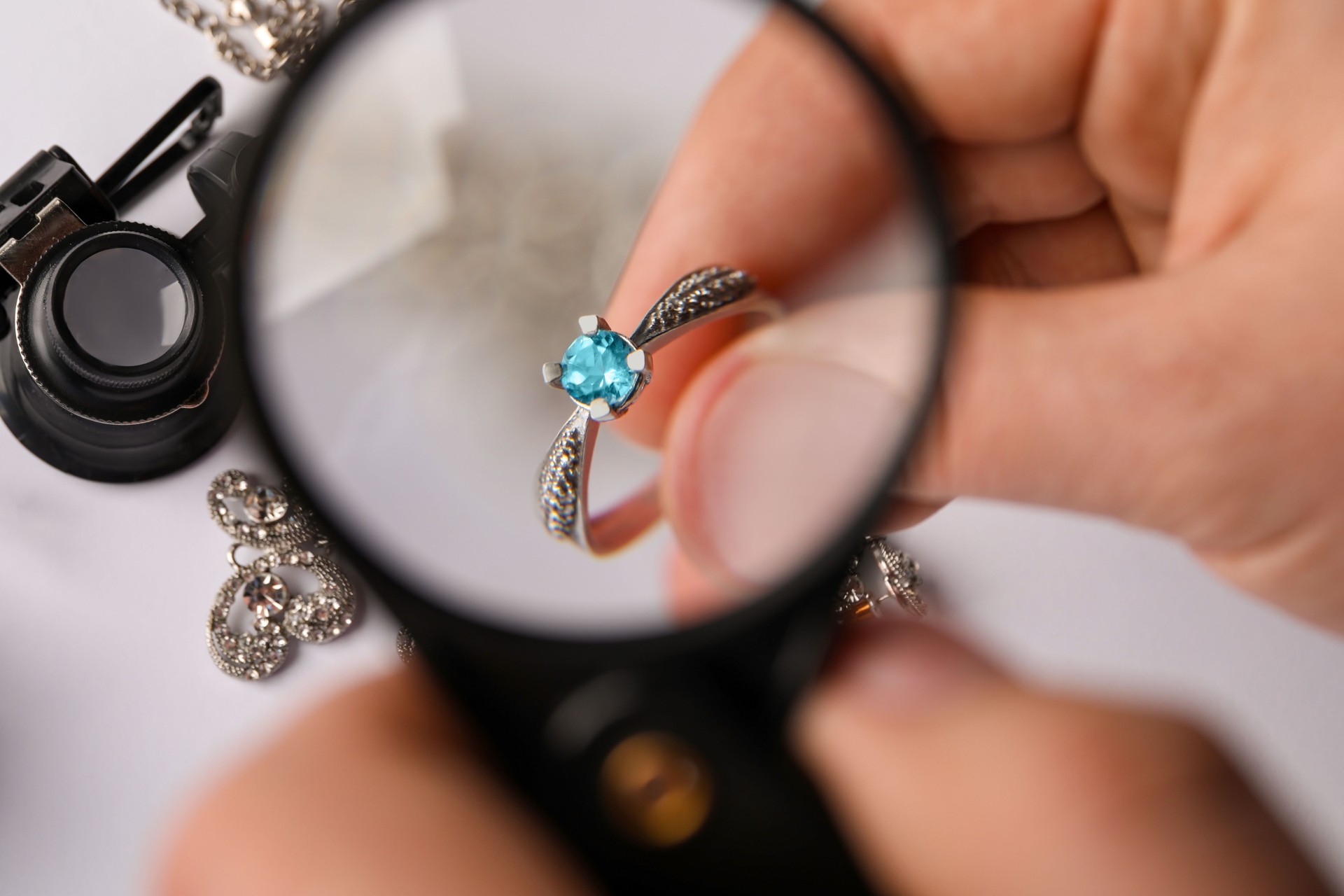 Jeweler examining topaz ring with magnifying glass at white table, closeup
